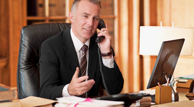A make attorney talking on a corded phone while seated at a desk covered in papers and folders