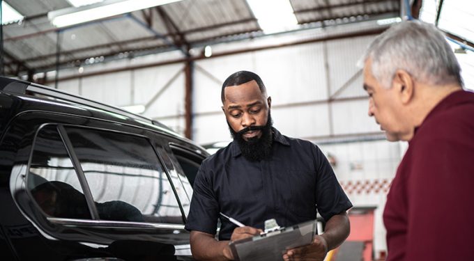 A bearded male auto service technician discussing a clipboard checklist with an elderly male customer