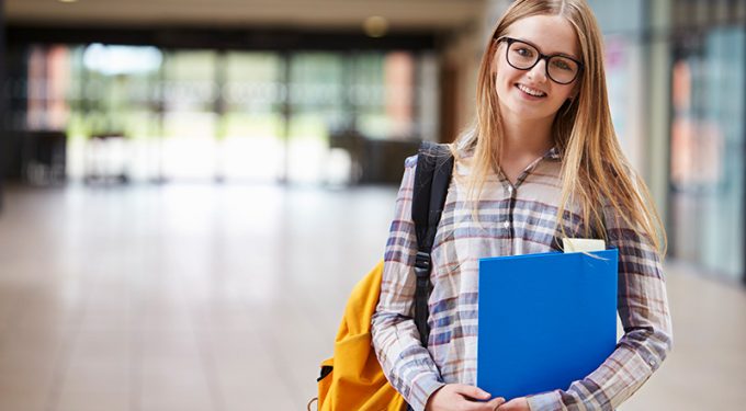 A young female student with long hair and glasses, holding a blue folder and shouldering a bag while standing in an empty hall of a school